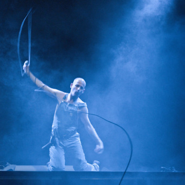 Eine Szene aus Work Body: Das Foto zeigt Michael Turinsky in einer Latzhose auf der Bühne sitzen. Er hält seinen rechten Arm in die Luft, in der Hand hält er ein Mikrofon, das verkabelt ist. Das Mikrokabel schwingt in der Luft.