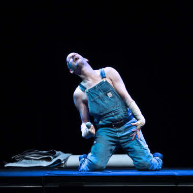 A scene from Work Body: The photo shows Michael Turinsky sitting on stage in dungarees. He throws his head back and holds a microphone in his hand. The stage is bathed in blue light. 