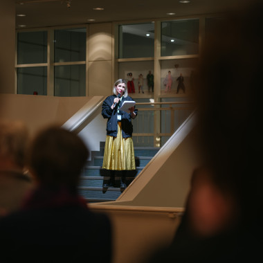 Festivaleröffnung im Foyer des Schauspielhaus Hannover. Das Foto zeigt Melanie Zimmermann bei der Eröffnungsrede. Sie steht auf einer Treppe und spricht in das Mikrofon in der Hand. Im Bildvordergrund das Festivalpublikum von hinten.