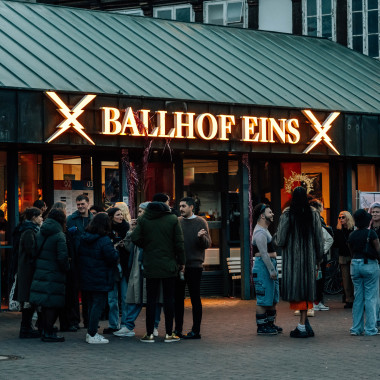 Ein Foto von der Spielstätte Ballhof Eins von außen. Es zeigt mehrere Menschen vor dem Eingang stehen. Über dem Eingang die leuchtende Schrift "Ballhof Eins".