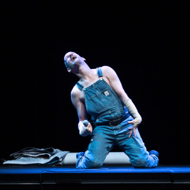 Eine Szene aus Work Body: Das Foto zeigt Michael Turinsky in einer Latzhose auf der Bühne sitzen. Er wirft den Kopf nach hinten und hält ein Mikrofon in seiner Hand. Die Bühne ist in blaues Licht getaucht. 